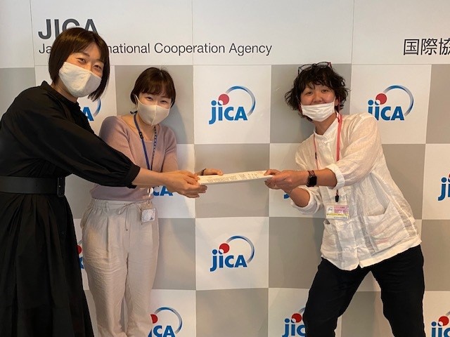 JICA東京にて。JICAと書いてある壁を背景に、廣瀬さんがJICA東京の担当スタッフ・小林様、篠崎様に契約書を手渡しているところ。